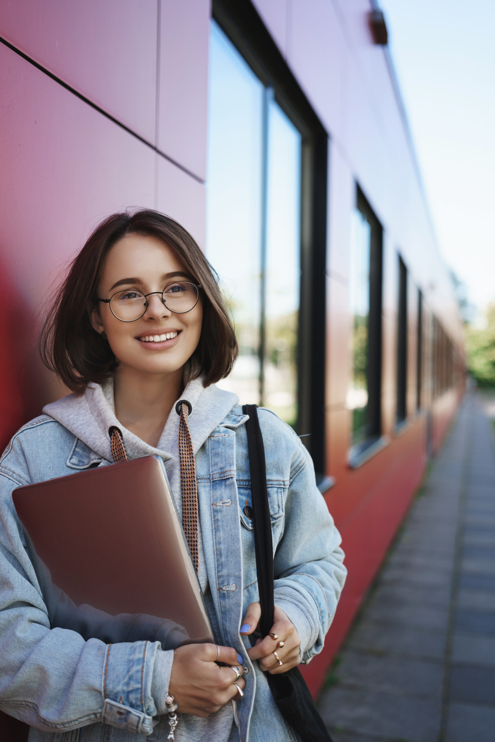 Vertical portrait of happy dreamy young female student look away with pleased expression, smiling, holding laptop on her way back to dormitory or work, enjoying spring sunny day, study with friend.